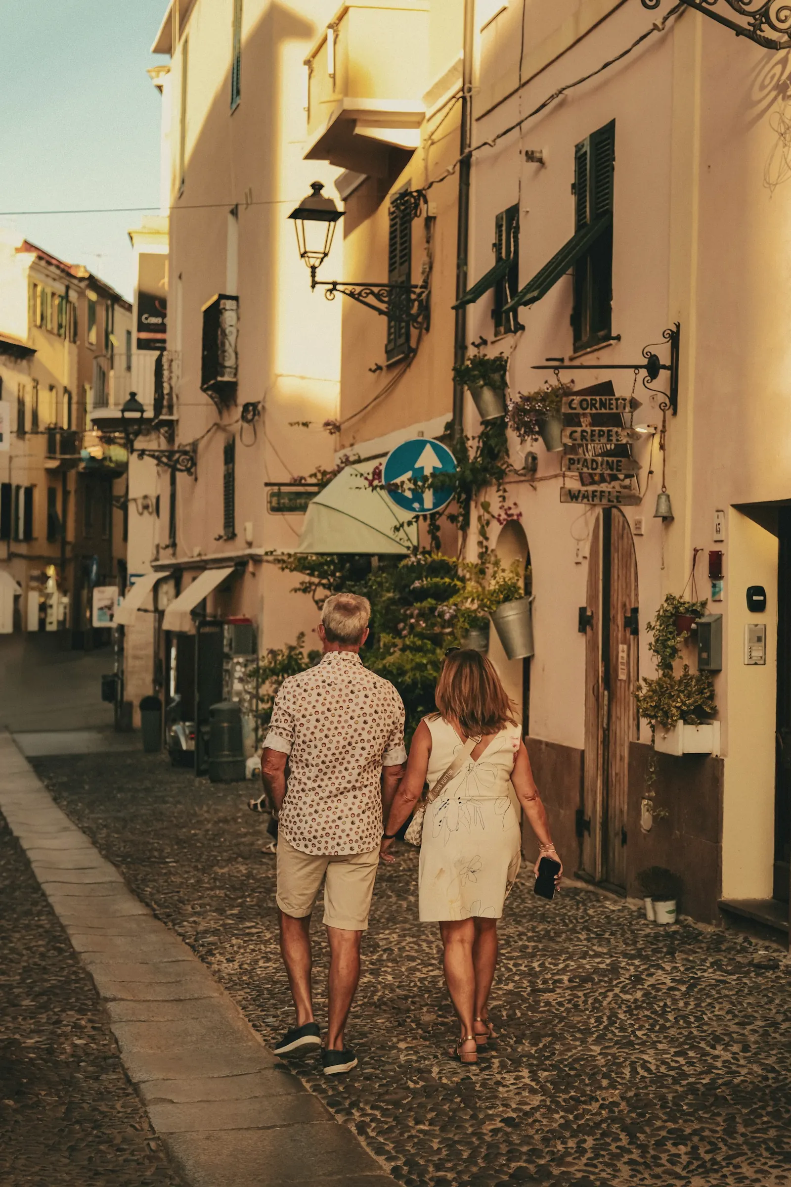 A couple holding hands, walking up a sunlit lane in an Italian town