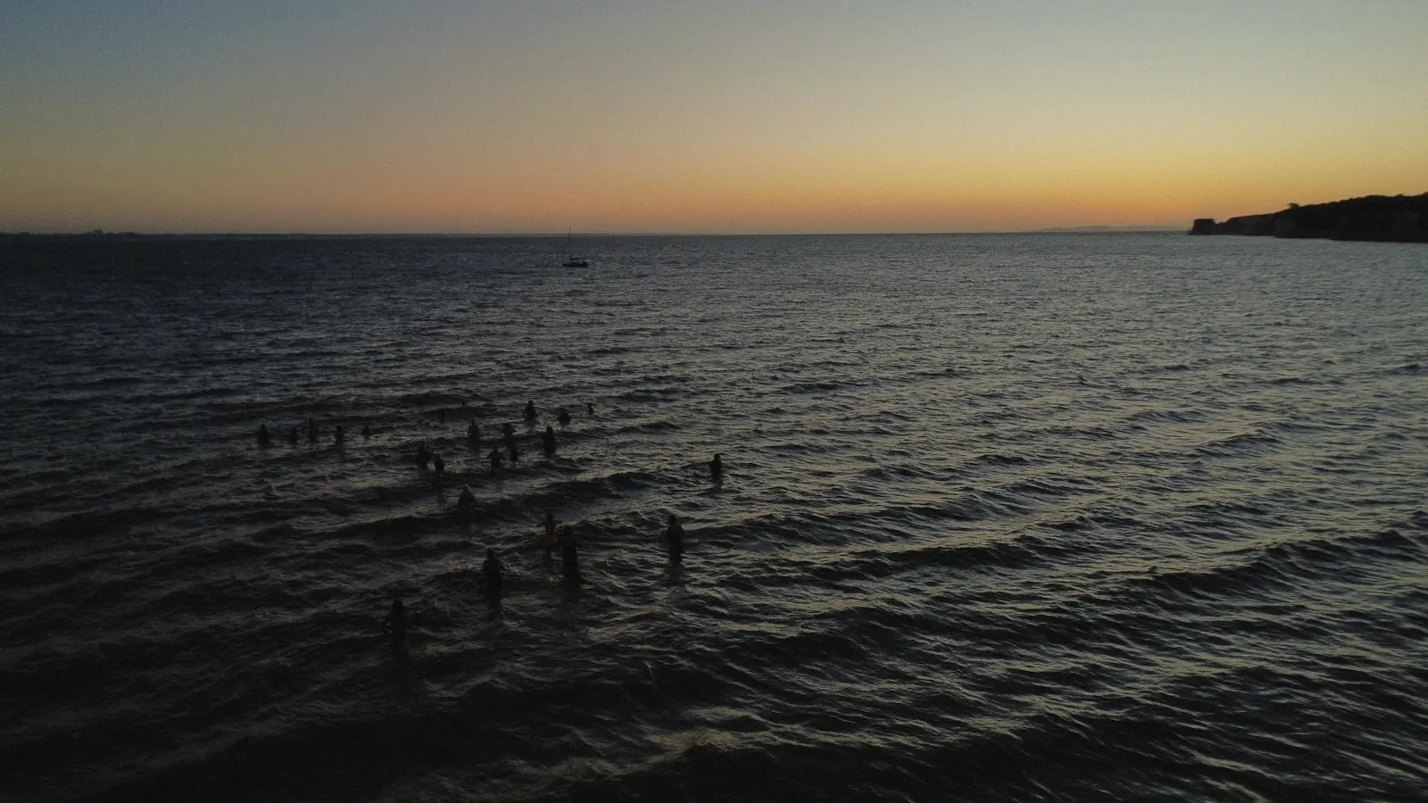 Swimmers in a calm sea at dusk, the sky glowing orange on the horizon