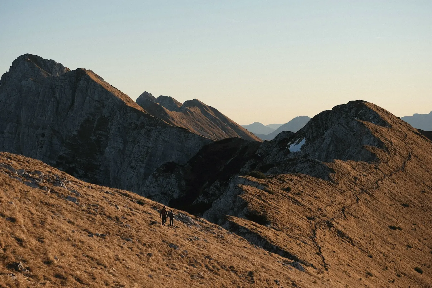 Two hikers small on a sunlit mountain ridge, jagged peaks beyond
