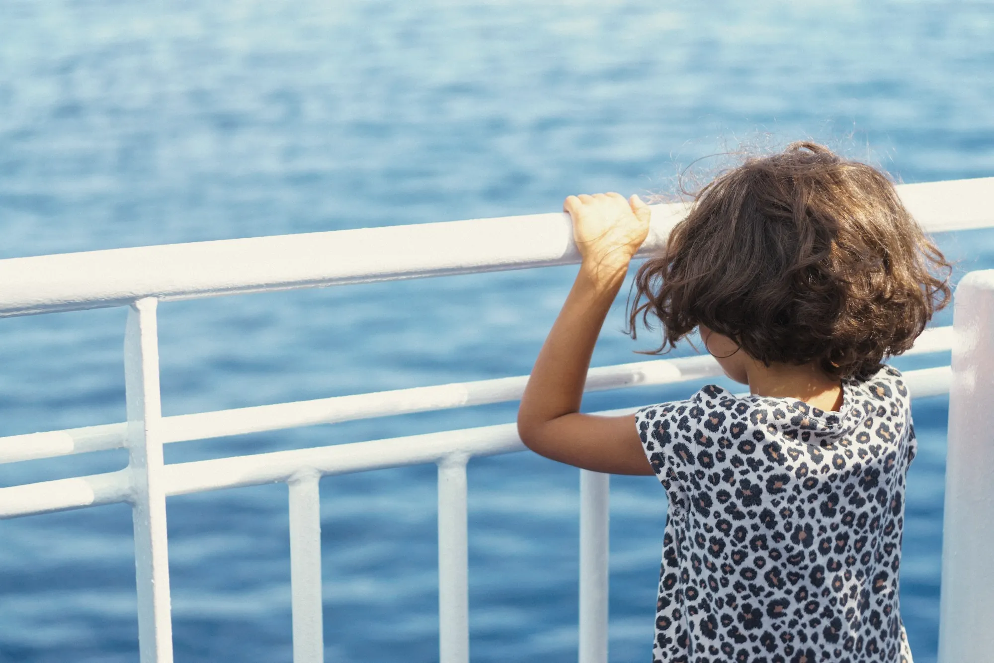 A young girl holding a ferry railing, looking out over a blue sea