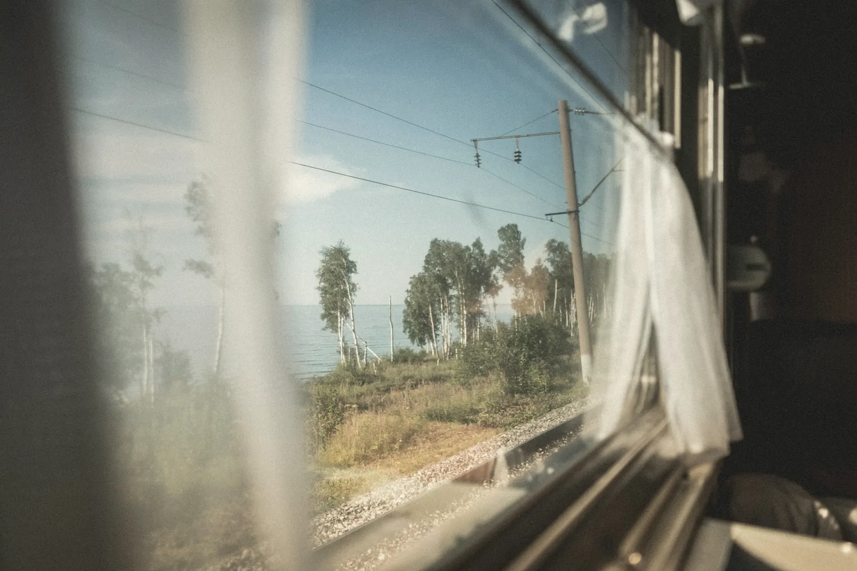 A train window with a white curtain lifting in the breeze, coastline passing outside